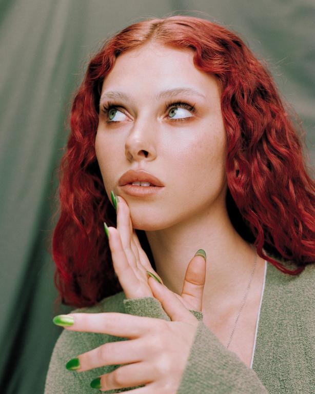 A woman with red hair and green nails looks up and to the right.