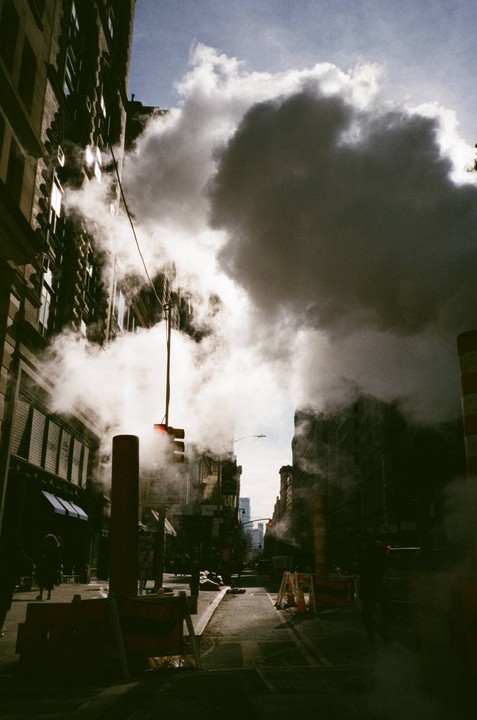 Steam billows from a street in a city with tall buildings under a cloudy sky.