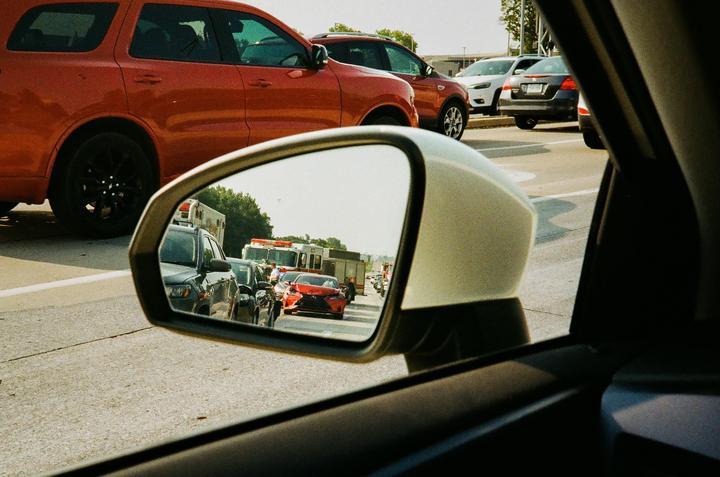 A car's side mirror reflects a traffic jam with emergency vehicles and a damaged red car.
