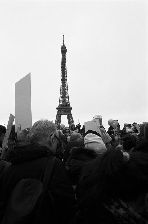 A large crowd of people stands in front of the Eiffel Tower, with some holding signs.