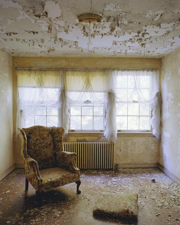 An abandoned room with peeling paint on the walls and ceiling contains a floral armchair, a footrest, and three windows with sheer white curtains.