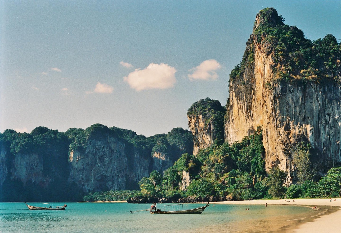 Two boats float in the turquoise water of a bay with a sandy beach and large, rocky cliffs covered in lush green foliage.