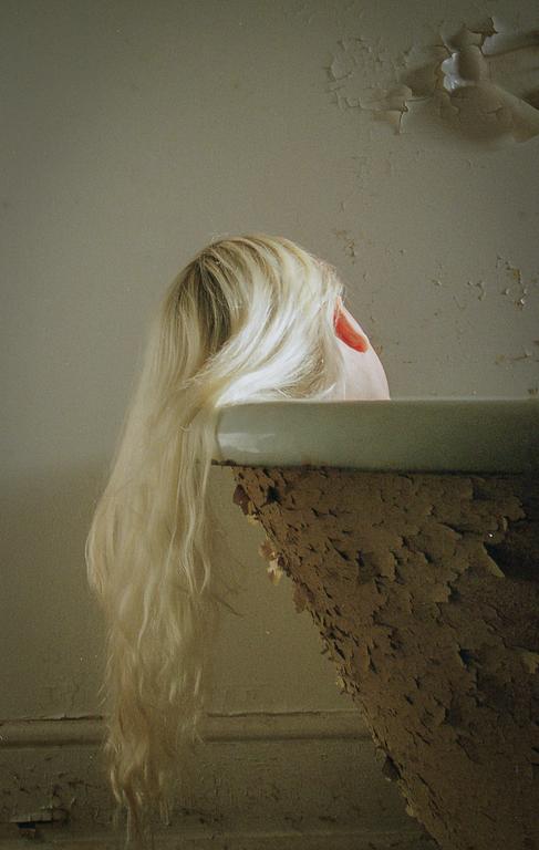 Blonde hair hangs over the edge of a decaying bathtub in a room with peeling paint.