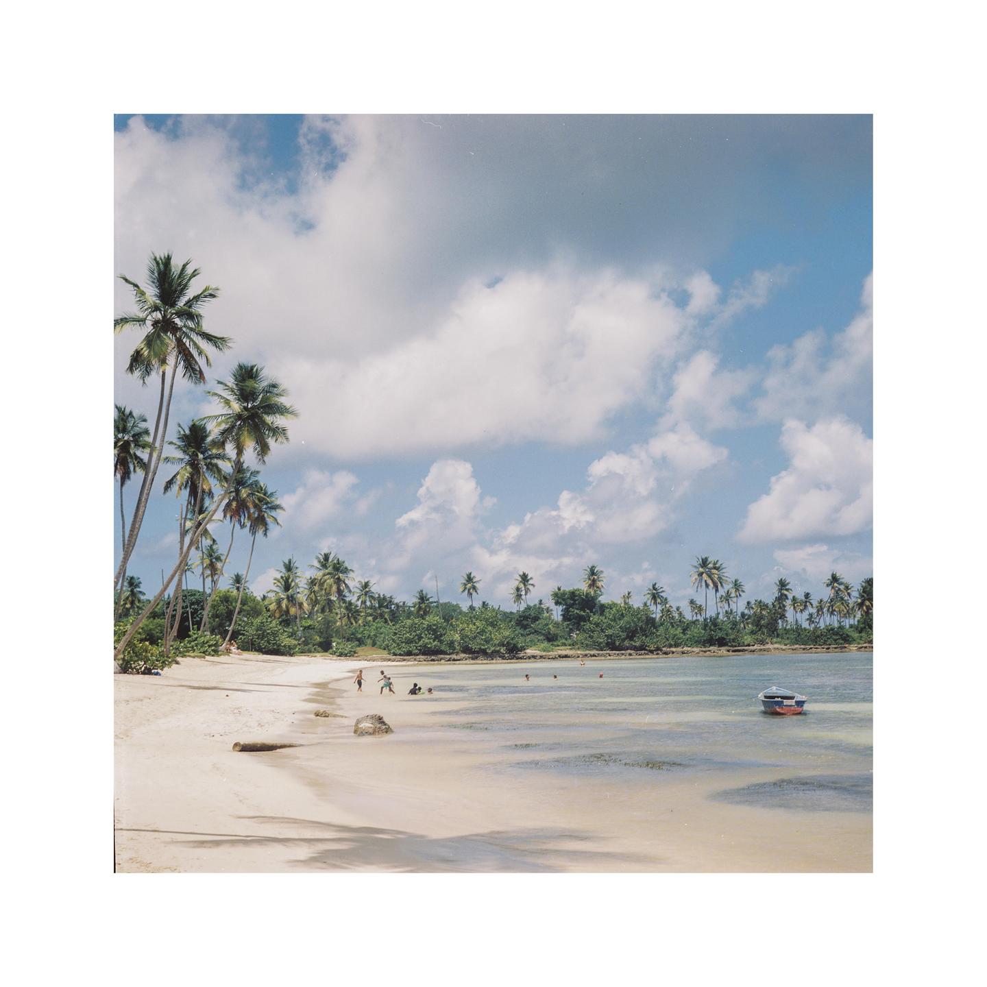 A sandy beach with palm trees and a small boat in the water under a cloudy sky.