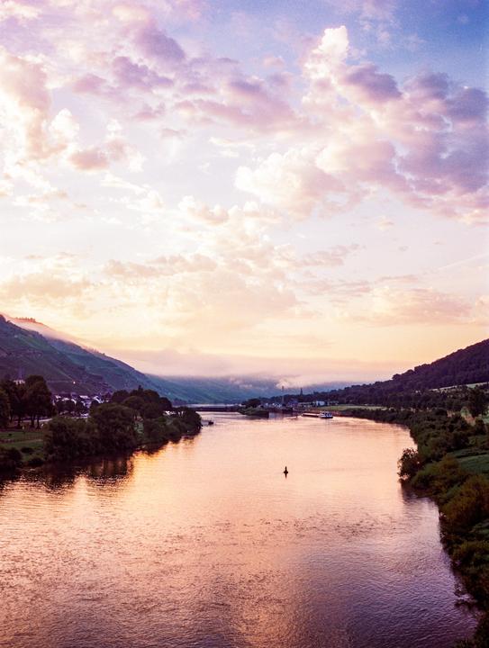 A wide river flows through a valley with hills on either side, under a sky filled with clouds at sunset.