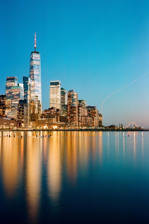 A cityscape with illuminated buildings reflects on the water at dusk.