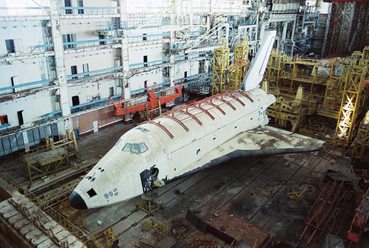 A large, white, abandoned space shuttle sits in a hangar at the Baikonur Cosmodrome.