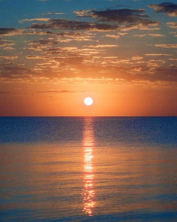 The sun sets over the ocean, casting a reflection on the water.