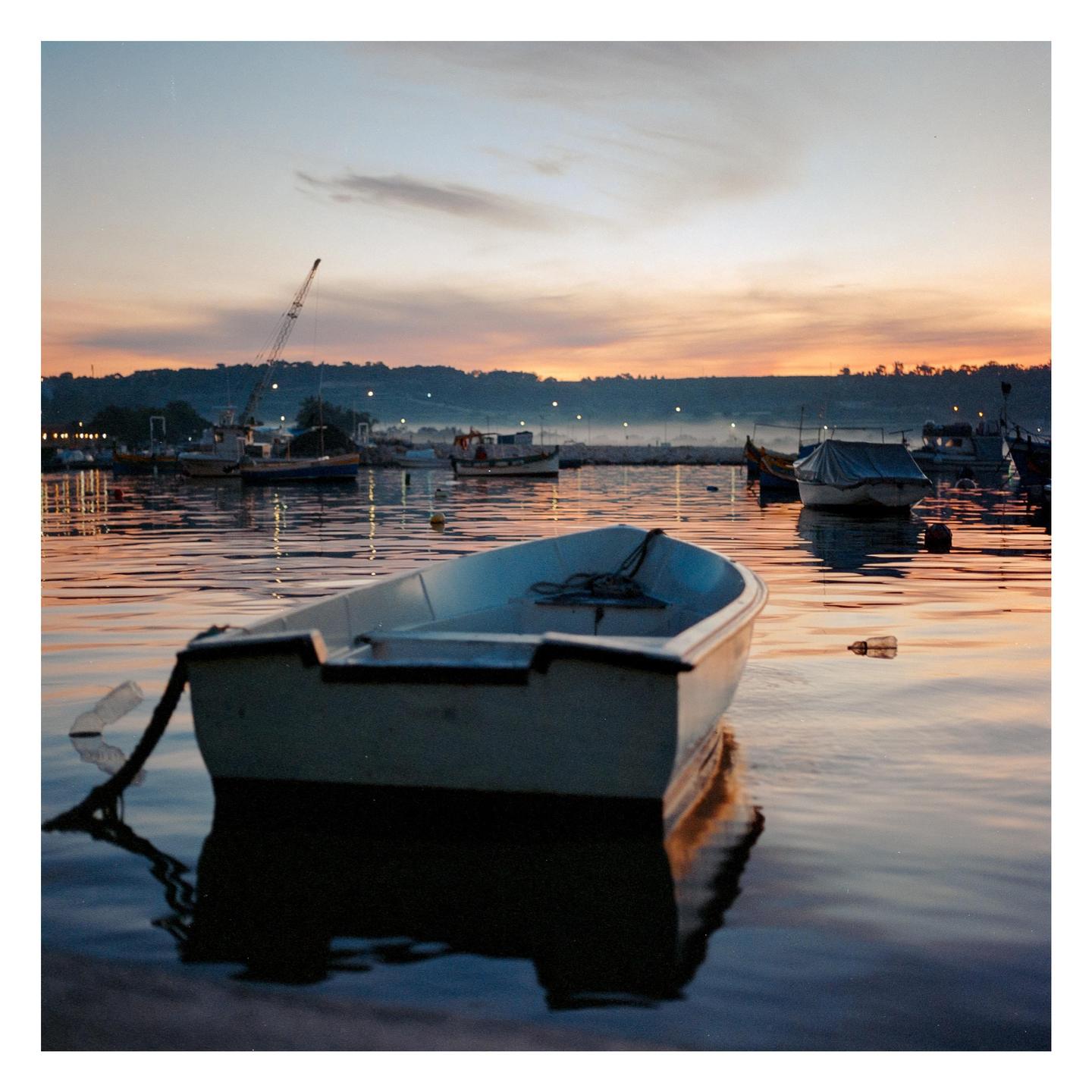 A small boat floats on the water at dawn, with other boats and a misty shoreline in the background.