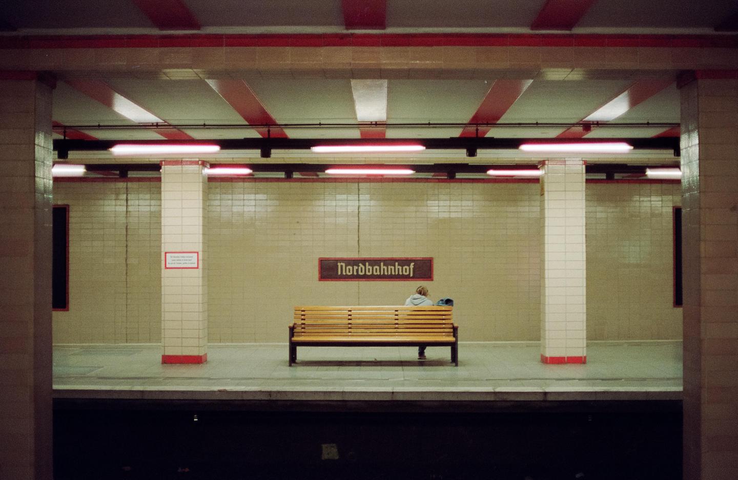 A person sits on a wooden bench in a subway station with a "Nordbahnhof" sign on the wall.