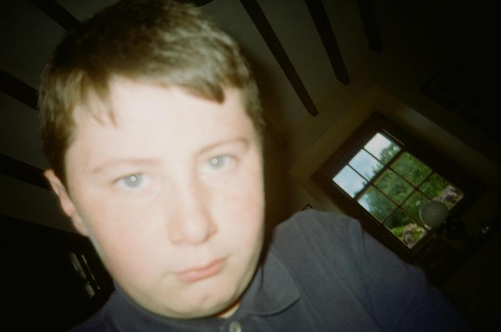 A boy with light brown hair and blue eyes looks directly at the camera, with a window showing green trees in the background.