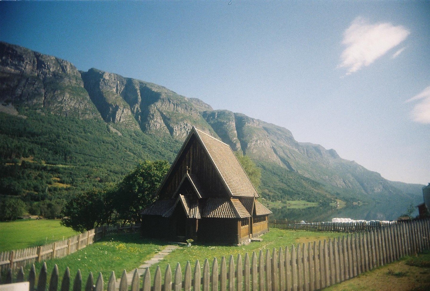 A traditional wooden church with a steep roof stands in a grassy field with a wooden fence in the foreground, backed by a large, tree-covered mountain range and a calm lake under a clear sky.