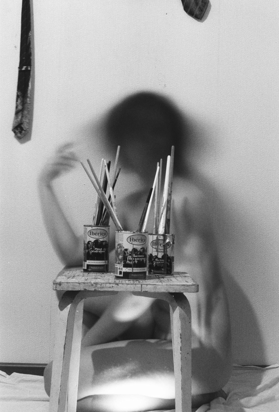 A blurred figure sits behind a stool holding three cans filled with paintbrushes, with a shadow cast on the wall.