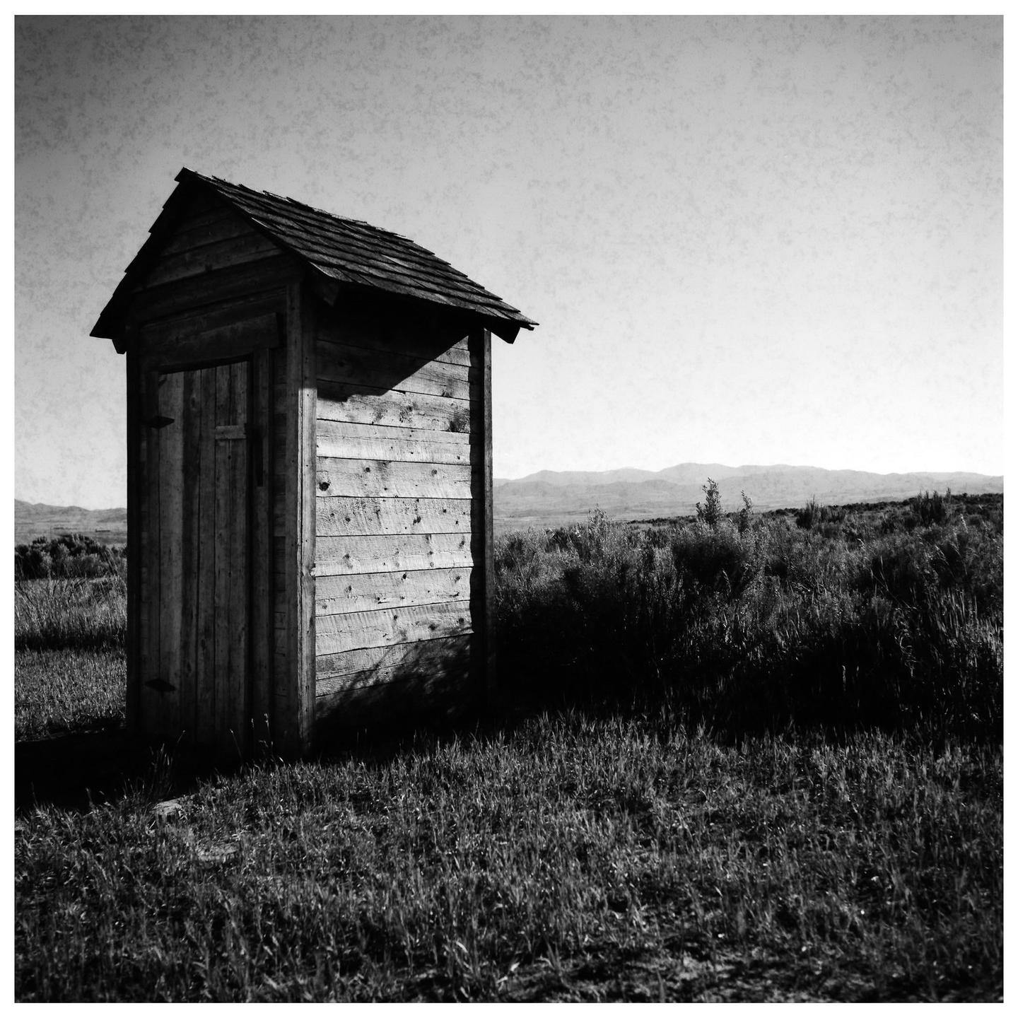 A wooden outhouse stands in a grassy field with mountains in the distance.