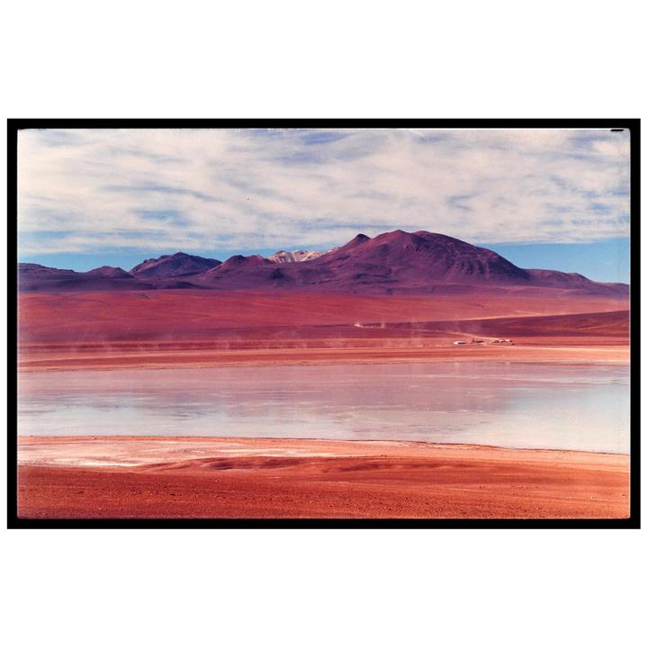 A vast landscape features a body of water reflecting the sky, with red mountains and a cloudy sky in the background.
