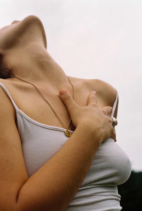 A woman wearing a white tank top and a gold necklace is looking up with her hand on her chest.
