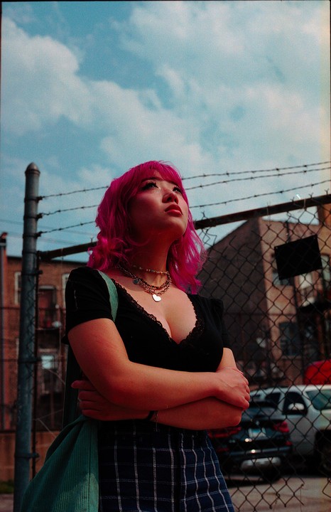 A woman with bright pink hair looks up at the sky, standing in front of a chain-link fence with barbed wire.