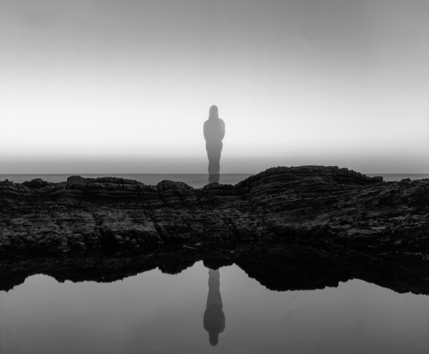 A ghostly figure stands in the water behind a rocky shore, with its reflection visible in a pool of water in the foreground.