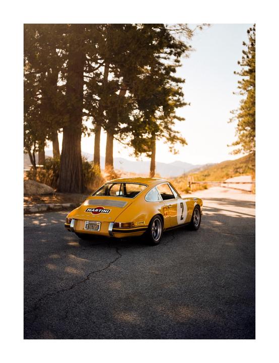 A yellow vintage sports car is parked on an asphalt road surrounded by tall trees and mountains in the distance.