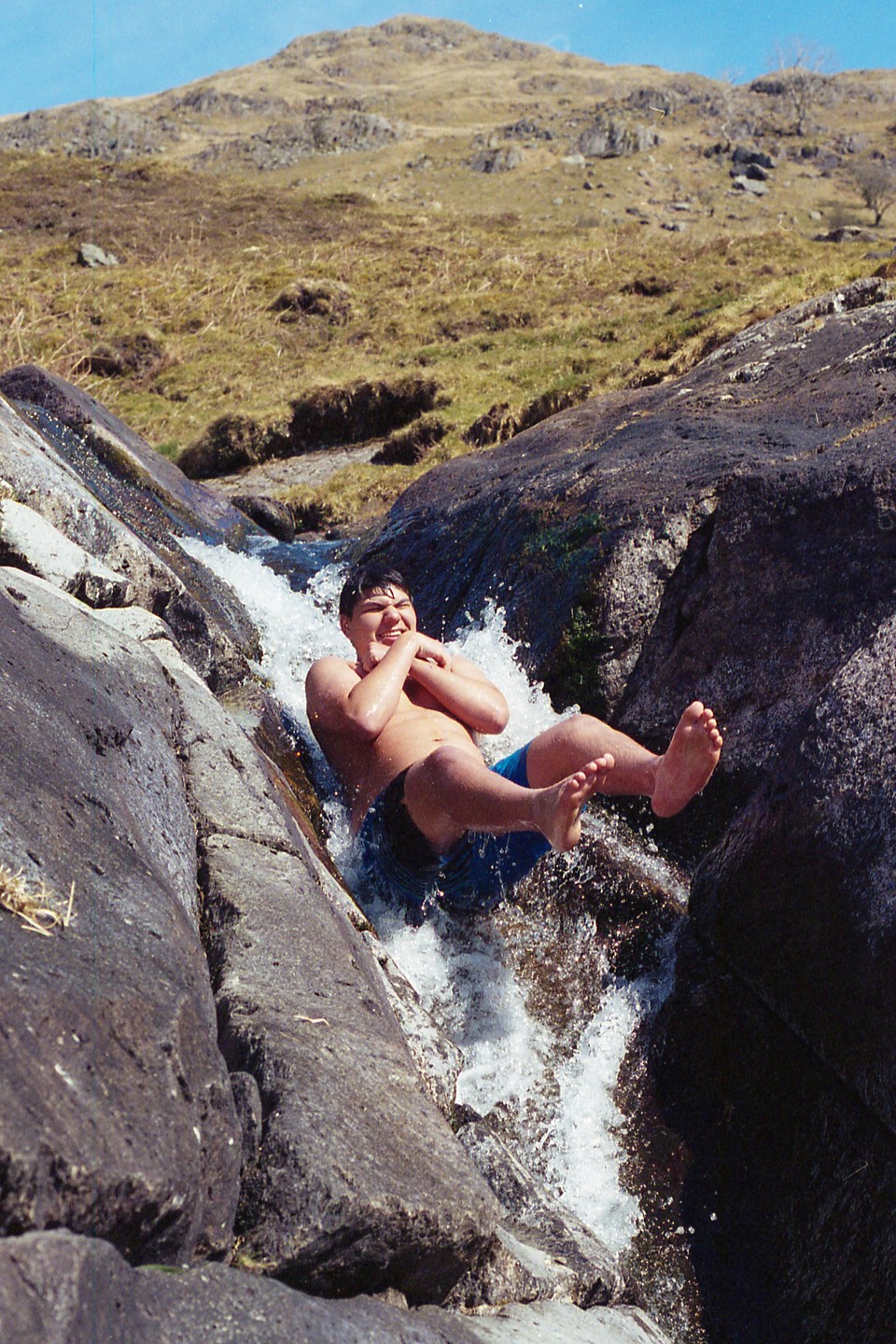 A young man slides down a natural rock slide into a pool of water in a mountainous landscape.