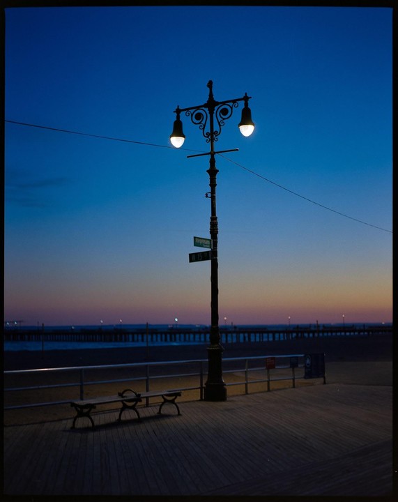 A street light illuminates a boardwalk at dusk with a pier and ocean in the background.