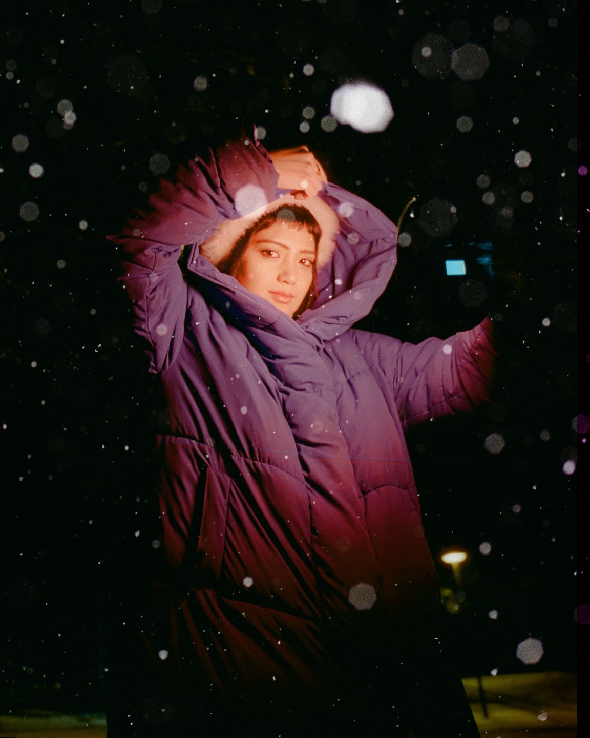 A woman in a purple winter coat stands in the snow at night.
