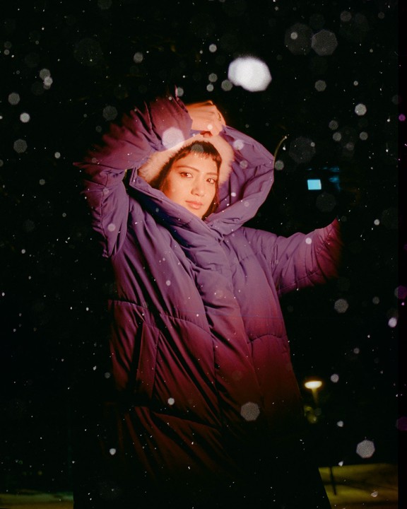 A woman in a purple winter coat stands in the snow at night.