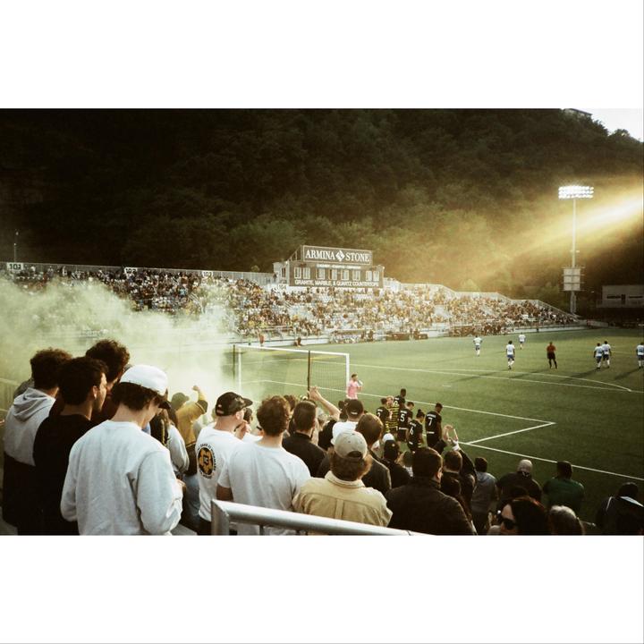 A crowd of people watches a soccer game in a stadium with a hillside in the background.