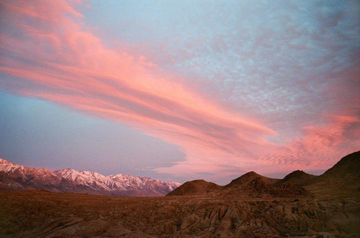 A desert landscape with snow-capped mountains in the distance is visible under a sky filled with pink and blue clouds.