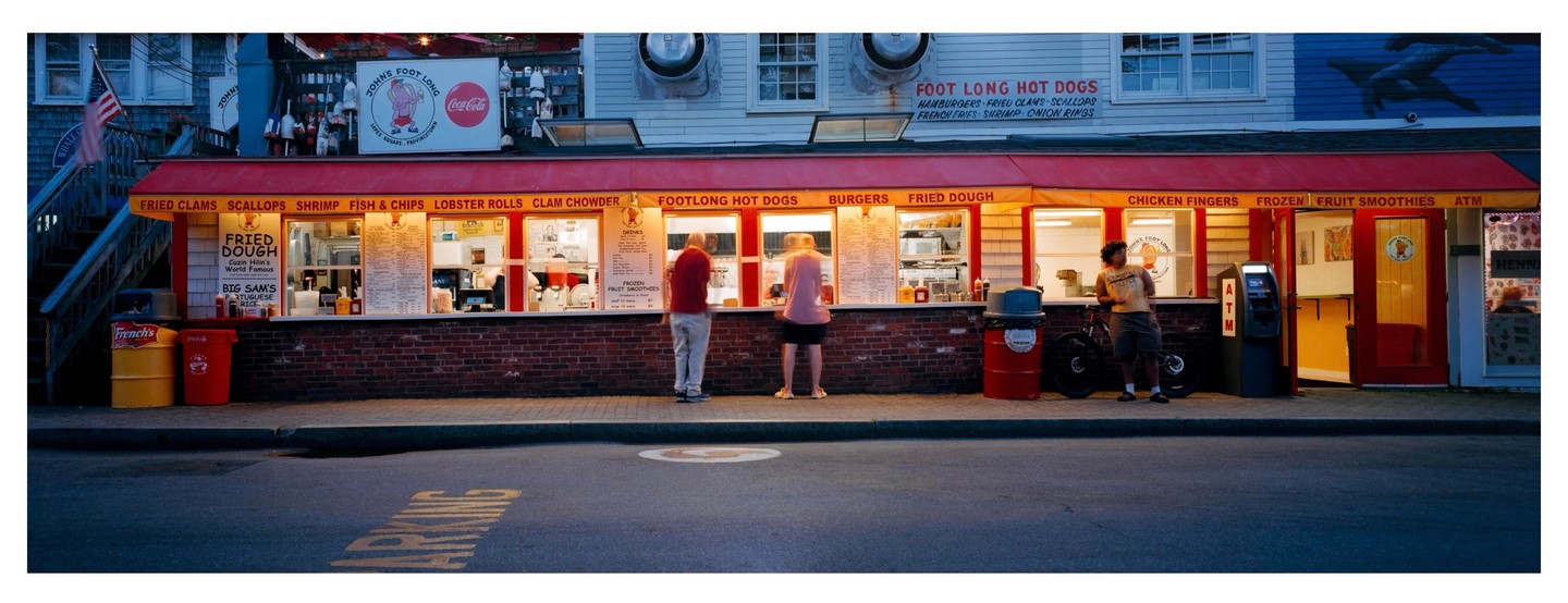 Three people stand in front of a brightly lit food stand with a red awning at dusk.
