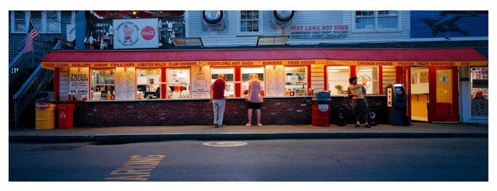 Three people stand in front of a brightly lit food stand with a red awning at dusk.