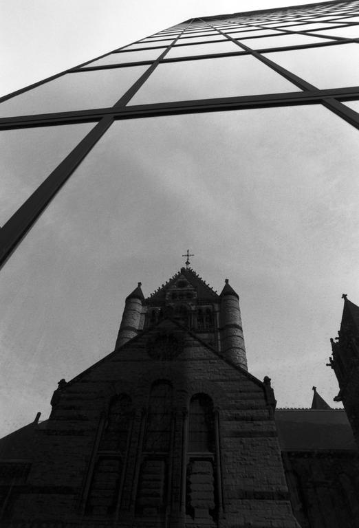 A church with a cross on its steeple is reflected in the glass facade of a modern skyscraper.