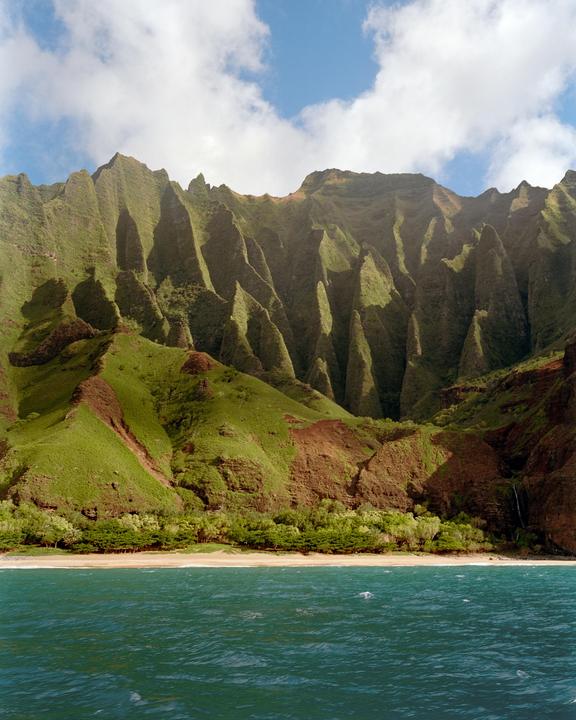 A lush green mountain range with sharp peaks rises above a sandy beach and turquoise ocean under a blue sky with white clouds.