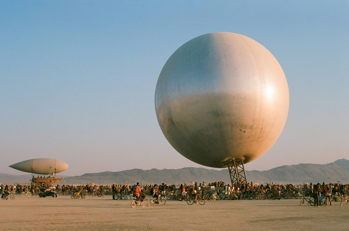 A large silver sphere and a blimp-like structure stand in a desert landscape filled with many people on bicycles.