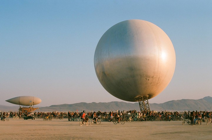 A large silver sphere and a blimp-like structure stand in a desert landscape filled with many people on bicycles.