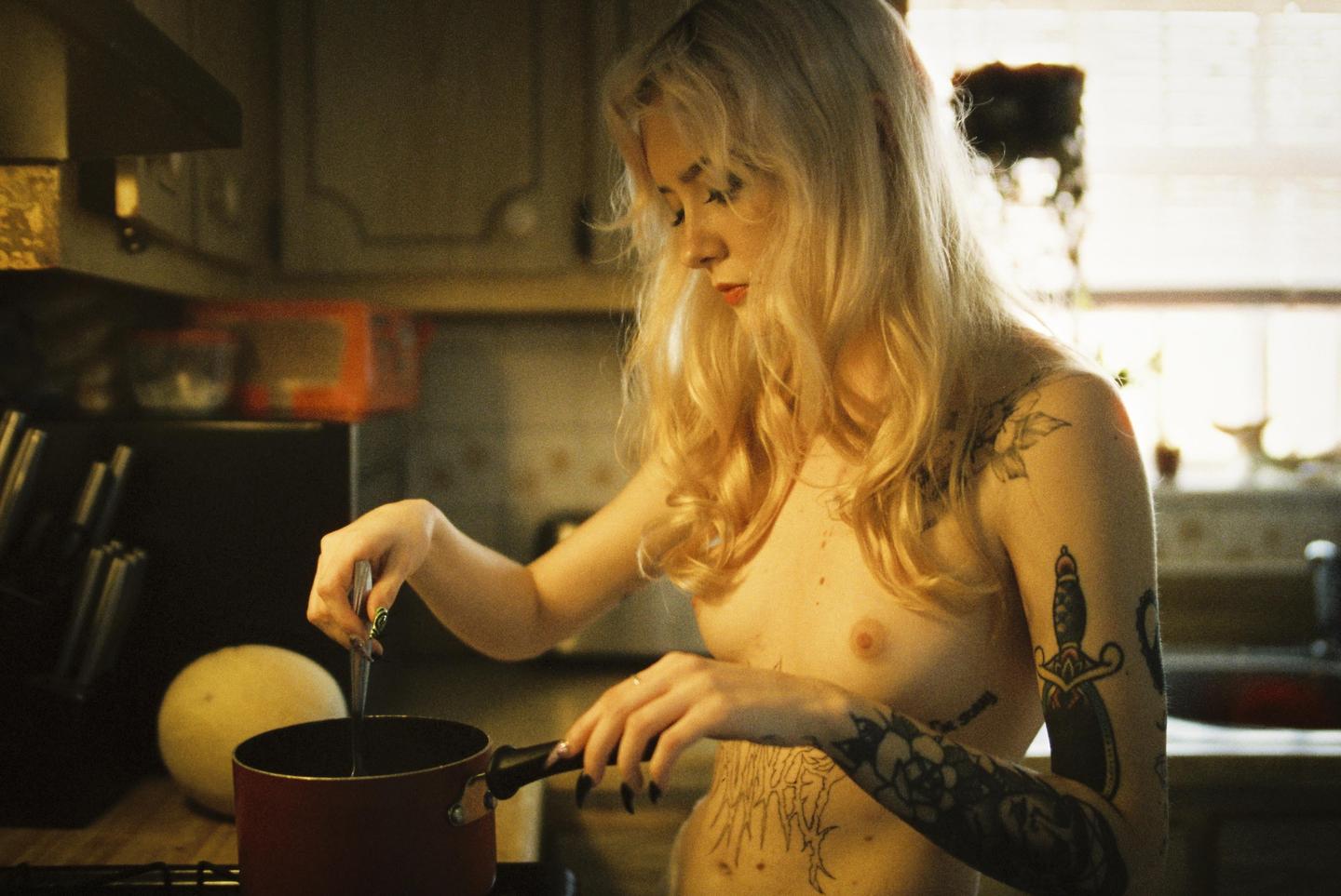 A woman with long blonde hair and tattoos on her arms and torso stirs a pot on a stove in a kitchen.
