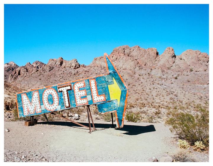 A weathered motel sign with an arrow points towards rocky desert mountains under a clear blue sky.