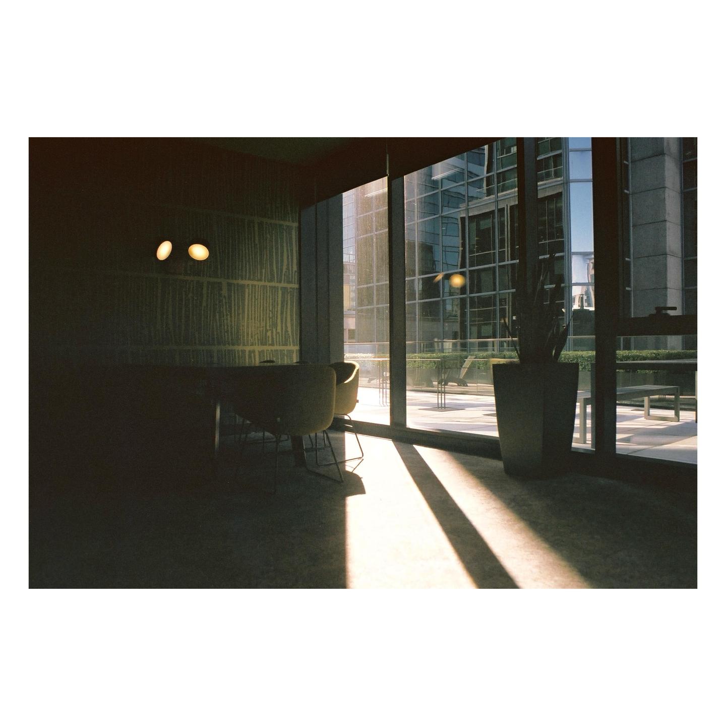 Sunlight streams through a large window into a room with chairs and a plant, revealing a modern building outside.