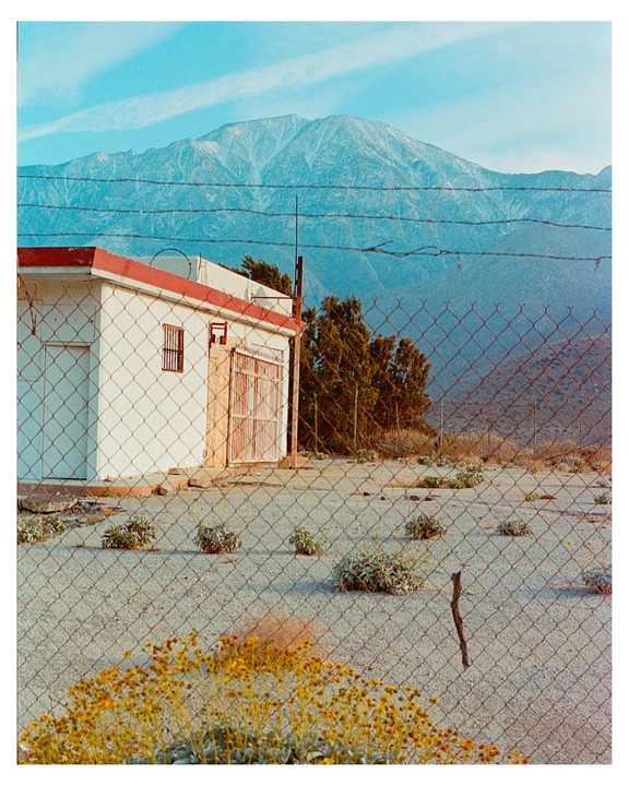 A white building with a red roof is visible through a chain link fence, with a large mountain in the background.