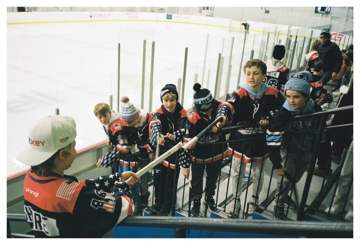 A group of boys in hockey jerseys and hats are gathered at the edge of an ice rink, with one boy holding a hockey stick.