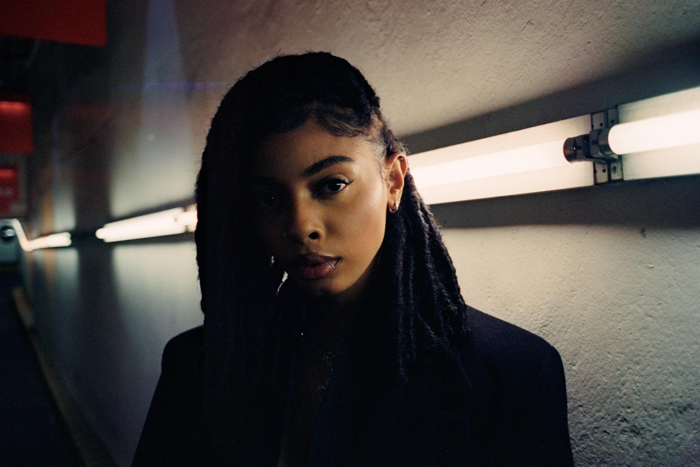 A woman with dreadlocks stands in a dimly lit tunnel with fluorescent lights on the wall.