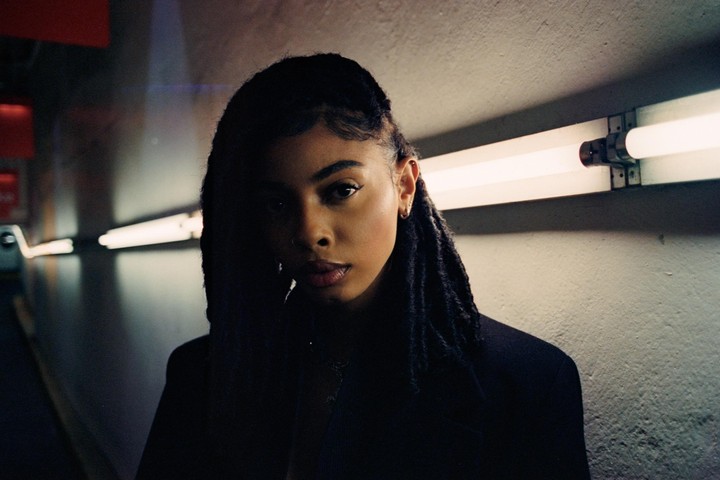 A woman with dreadlocks stands in a dimly lit tunnel with fluorescent lights on the wall.