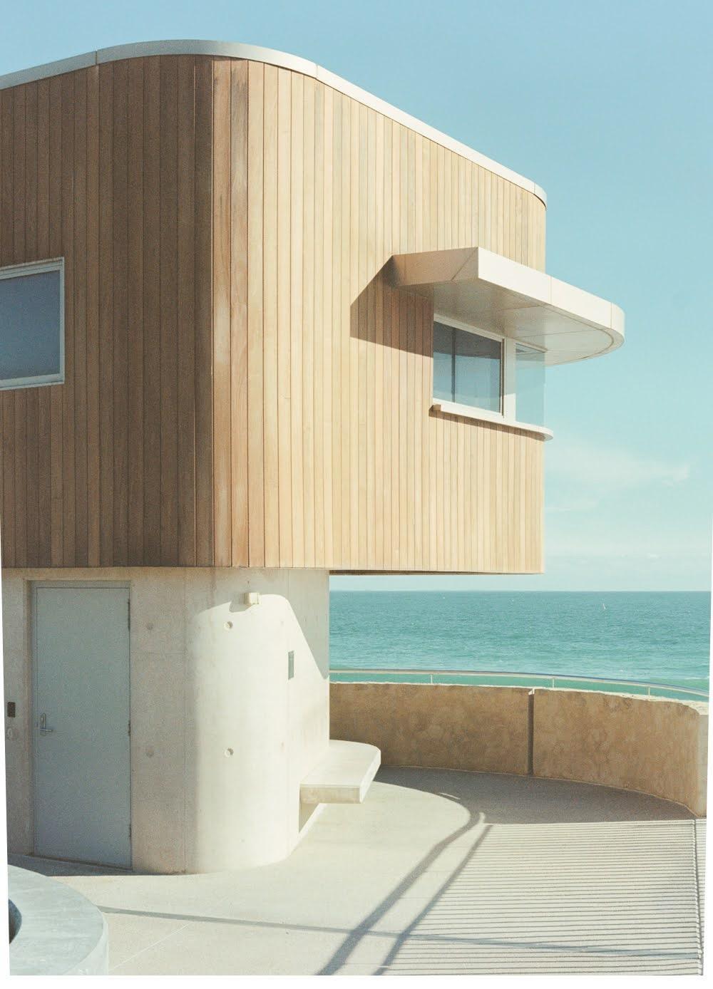 A modern building with wood siding and a concrete base overlooks the ocean.