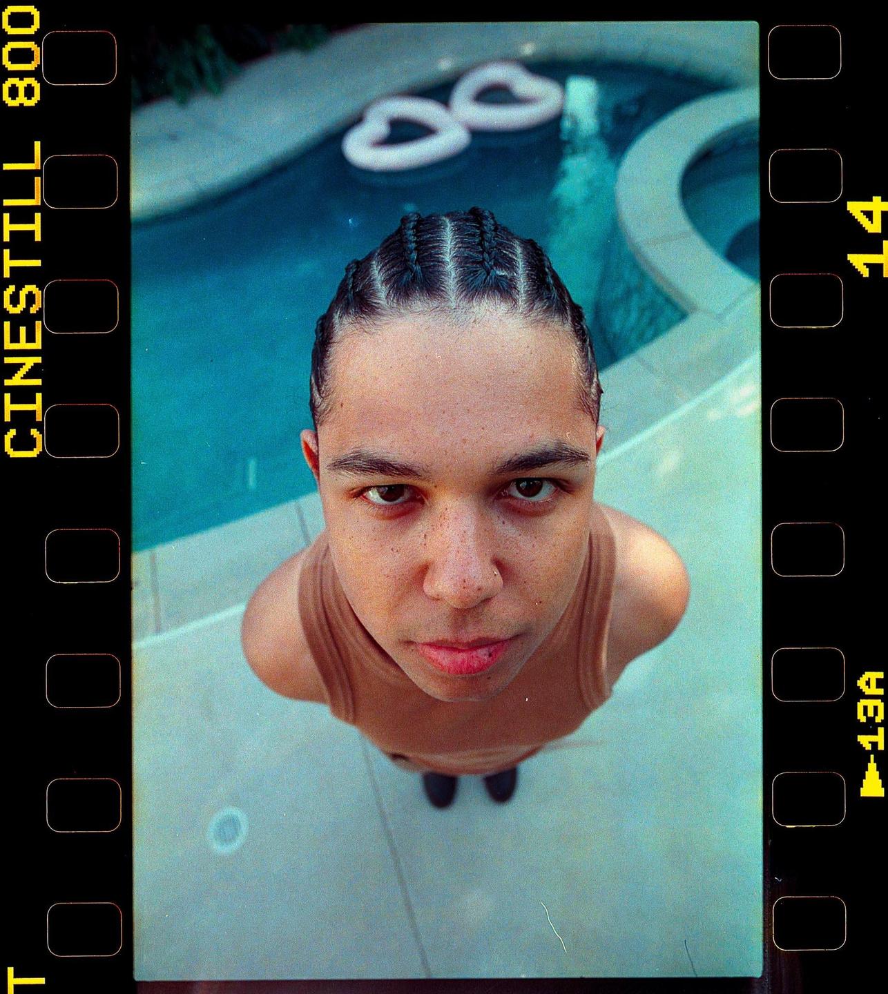 A man with braided hair and a tank top looks up at the camera from beside a swimming pool.