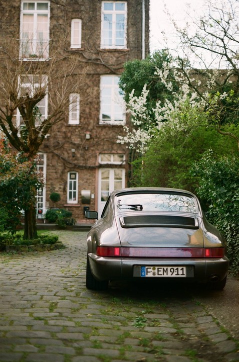 A gray Porsche 911 is parked on a cobblestone driveway in front of a brick building covered in ivy.
