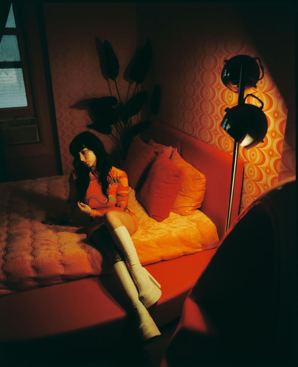 A woman in a patterned dress and white boots sits on a bed in a dimly lit room with patterned wallpaper.