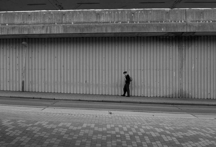 A woman walks along a sidewalk next to a corrugated wall under a concrete overpass.