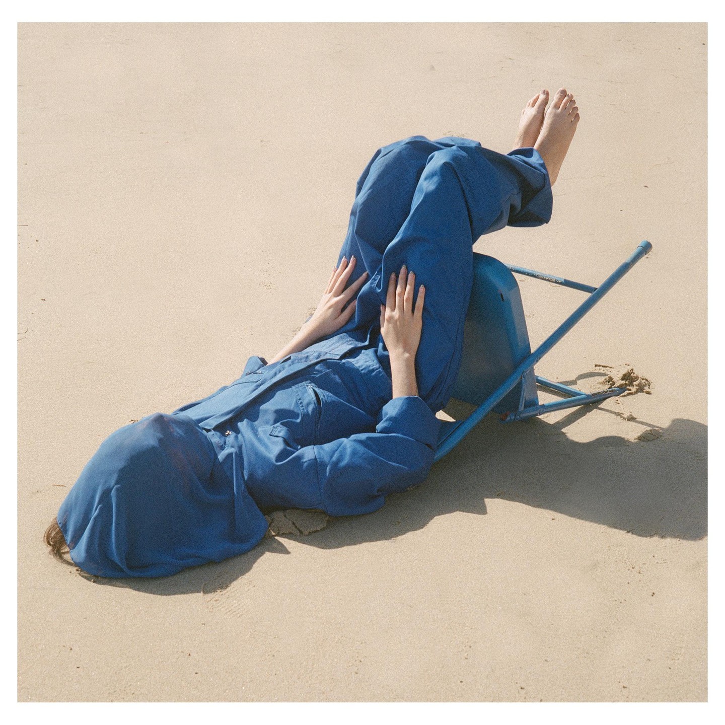 A person in a blue jumpsuit lies on a sandy beach with their head covered and legs propped up on an overturned blue chair.