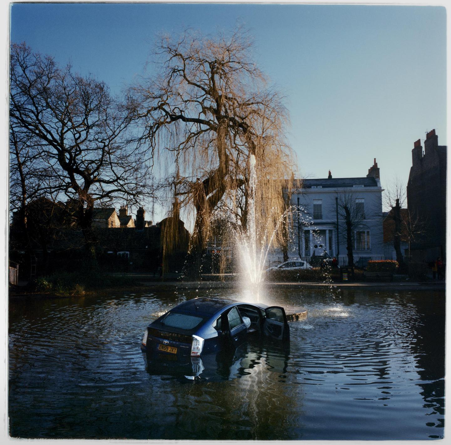 A blue car is partially submerged in a pond with a fountain spraying water next to it, surrounded by bare trees and buildings.