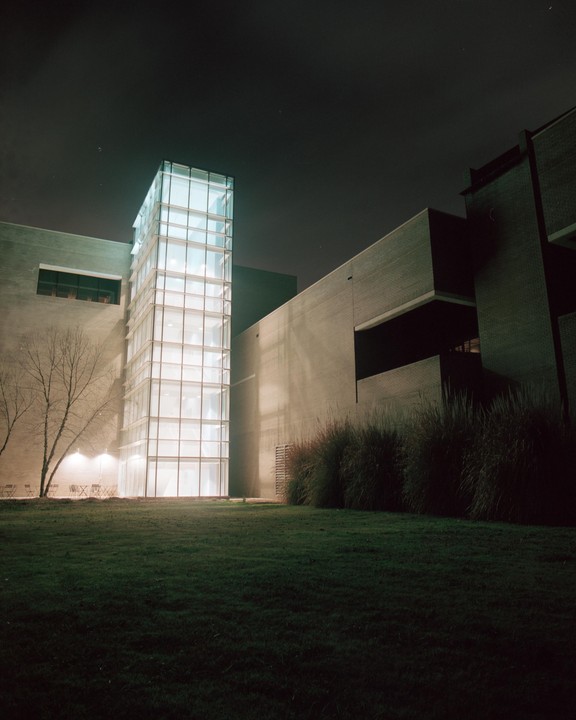 A modern building with a brightly lit glass tower stands next to a dark brick structure, with a grassy lawn and bare tree in the foreground at night.